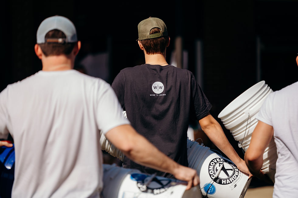 Two people carry white buckets outdoors. One wears a shirt with "Wine to Water" text. Bright sunlight highlights the scene.