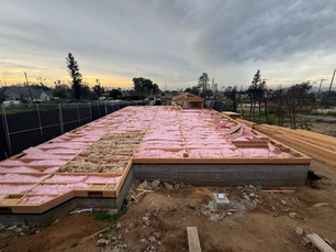 Construction site with pink insulation laid on wooden frame, surrounded by fences and trees under a cloudy sky at sunset.
