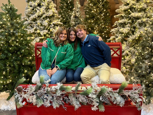 Three people, two girls one boy, sitting on a red bench surrounded by christmas greenery.