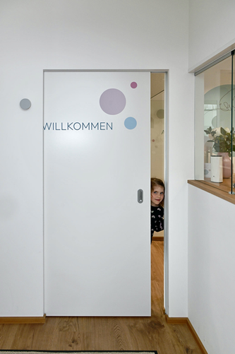 Kind schaut durch eine Tür, auf der Willkommen steht.