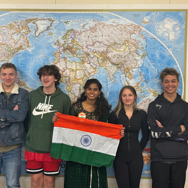 A photograph of students standing in front of a map of the world, holding up the flag of India.