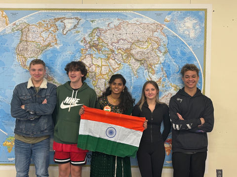 A photograph of students standing in front of a map of the world, holding up the flag of India.
