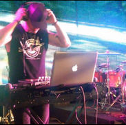 DJ Kolmega in a black sleeveless shirt and black pants on stage with turntables and laptop putting his headphones on.  There is a big giant digital screen in the background with special effects and in the background is also a drum kit.