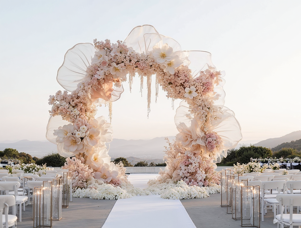Floral wedding arch with soft pink and white blooms, sheer fabric, and mountain view. Aisle lined with white chairs and lanterns.