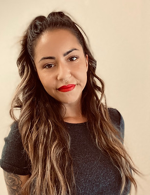 Porträt einer Frau südländischen Typs mit welligen Haaren und blonden Meches, leicht zusammengebunden. Sie trägt roten Lippenstift auf ihren vollen Lippen, dezentes Make-up mit leicht geschminkten Wimpern und mitteldicke, symmetrische Augenbrauen, die leicht nachgeschminkt sind. Ihre braunen Augen wirken freundlich, und sie lächelt leicht. Sie trägt ein graues Shirt, ihre Hände sind hinter dem Rücken verschränkt, und auf der rechten Seite ihres Oberarms ist ein Tattoo sichtba
