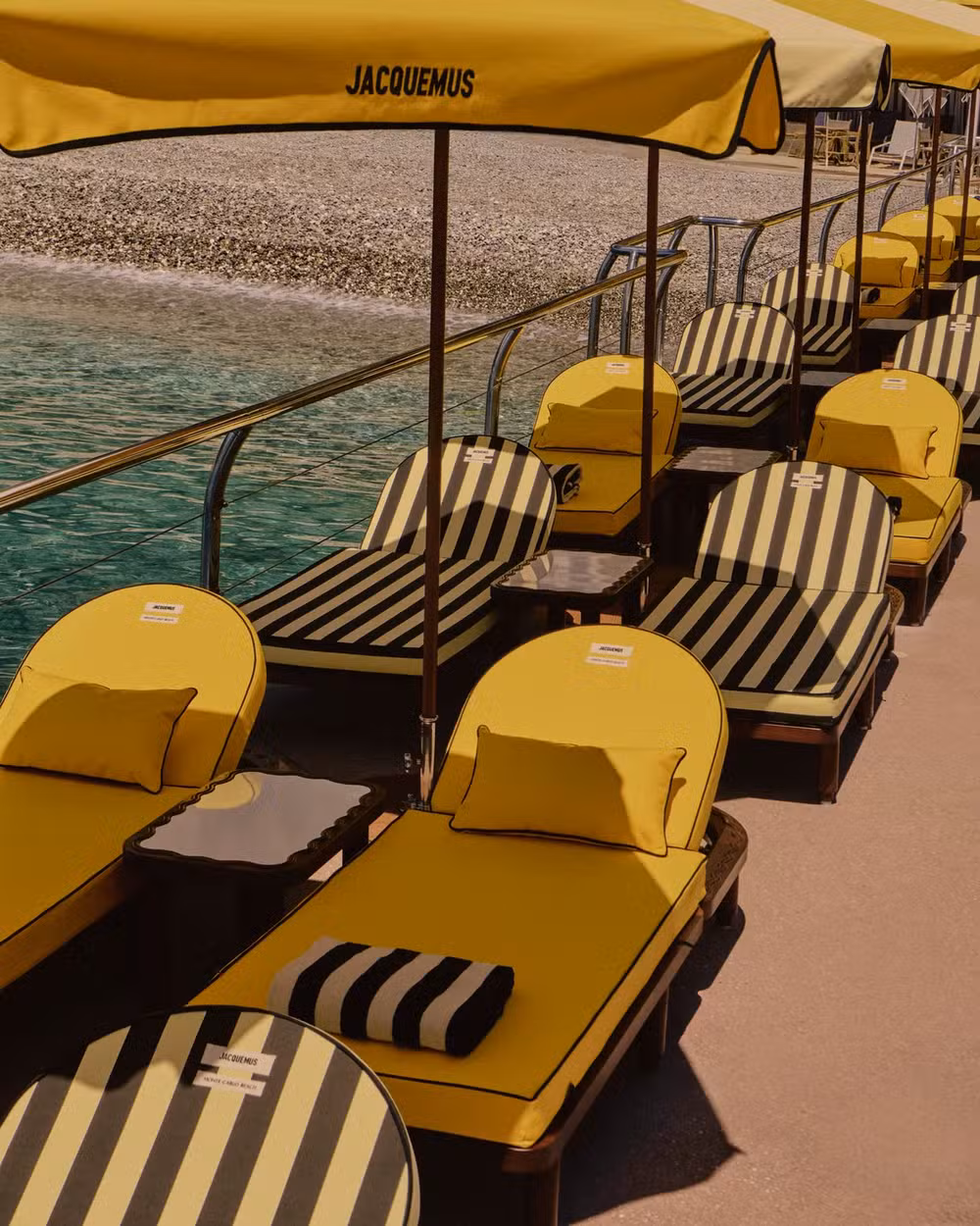 Espreguiçadeiras amarelas e guarda-sóis Jacquemus dispostos simetricamente sobre o píer do Monte-Carlo Beach, à beira do mar azul da Riviera Francesa.