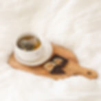 Xícara de chá em cerâmica branca sobre tábua de madeira com chocolate e cítricos secos, em cenário com tecido claro — ritual de pausa com suavidade e prazer sensorial.