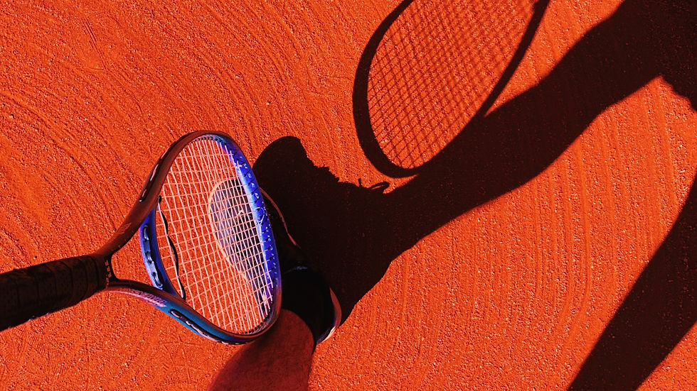 looking down on a clay court with a guy's legs and racket creating a shadow next to him