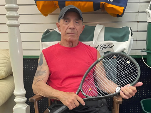 Barky Boodakian sitting in a tennis shop holding his racket, wearing a red sleeveless shirt and black cap. One of The Racket Doctor’s first and longest-standing customers since 1970.