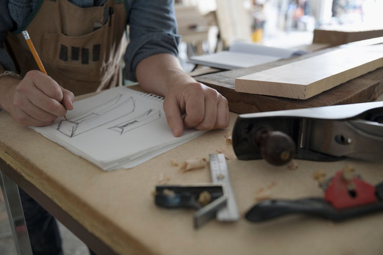 Bespoke carpenter sketching oak furniture design at woodworking bench with tools in workshop.