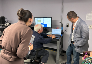 Three people analyzing data on a laptop in an office. A woman takes notes, a man points at the screen, and another observes. Monitors display hair claims testing graphs.