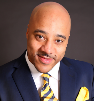 Ovi Kedo in a navy suit and yellow striped tie smiles warmly against a dark background, exuding confidence and friendliness.