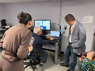 Three people gather around a desk, watching a man use a laptop and monitor in a lab, with graphs on screen.