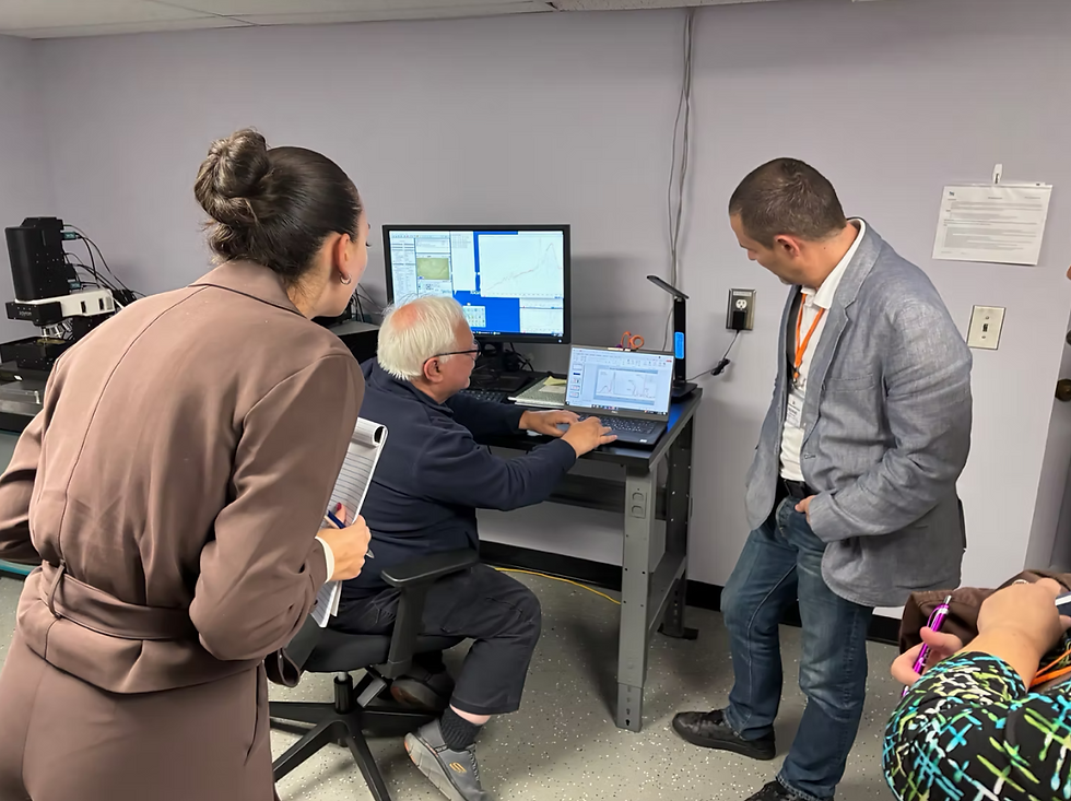 Three people gather around a desk, watching a man use a laptop and monitor in a lab, with graphs on screen.