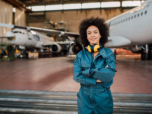 Woman in blue flight suit with yellow ear defenders stands smiling in an aircraft hangar beside two planes.