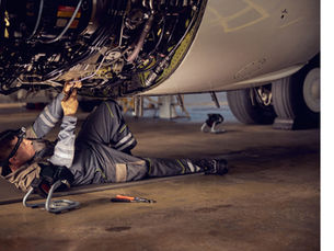 Aircraft mechanic in gray coveralls repairs wiring under a jet in a hangar, with tools on the floor.