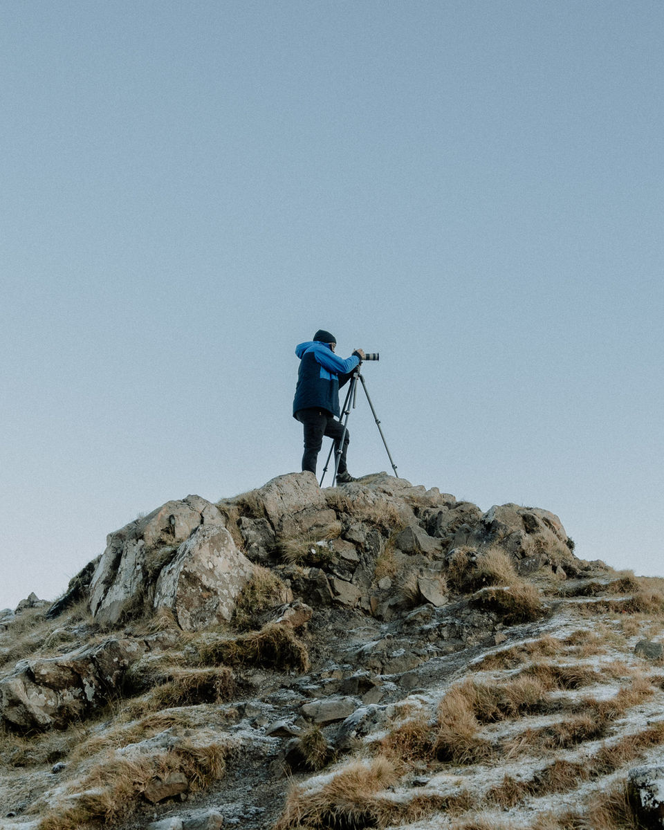 A lone photographer on a rocky outcrop under a pale blue sky.