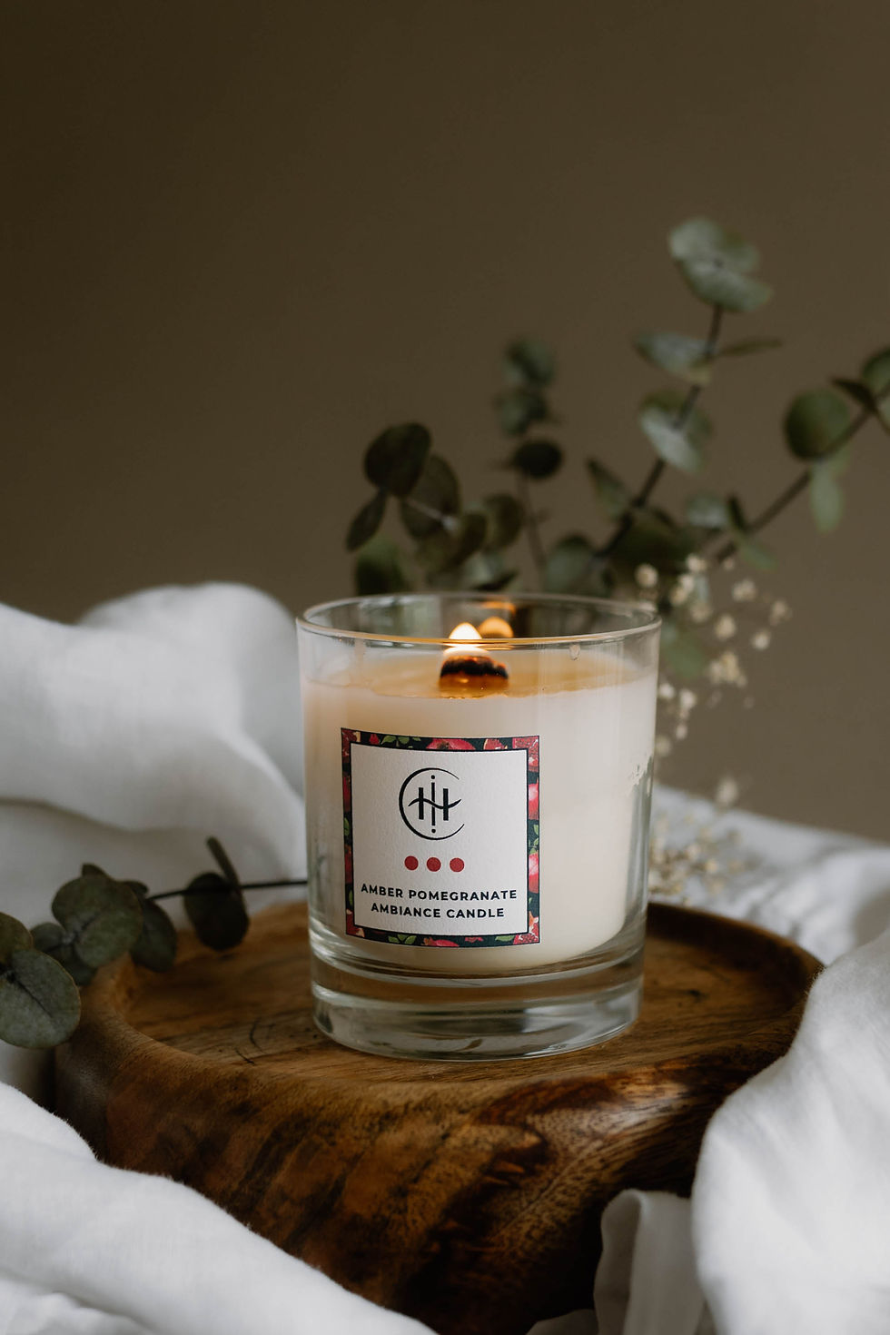Amber Pomegranate candle burning on a wooden tray with greenery and soft white cloth