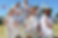 Girls in white cricket uniforms and sun hats on a field, smiling and holding cricket balls and bats under a clear blue sky.