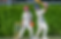 Two young cricket players in white, one batting with a blue helmet, the other as wicketkeeper in a red helmet, play on a green field.