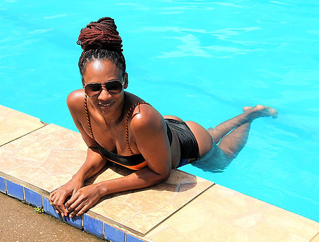Lachende Frau in Sonnenbrille und schwarzem Badeanzug am blauen Poolrand.