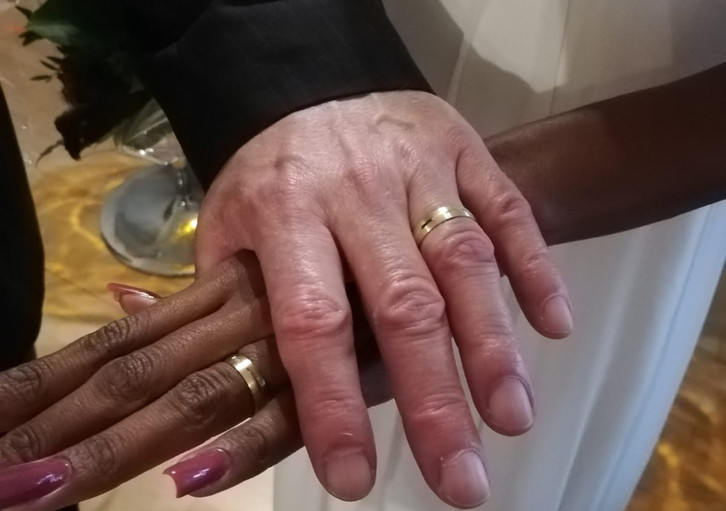 Zwei Hände verschiedener Hautfarben mit goldenen Eheringen, die sich halten.