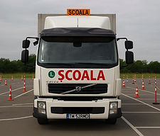ein weißer Volvo Lastwagen, der für Fahrschulzwecke genutzt wird. Auf der Motorhaube und über der Windschutzscheibe steht in großen roten Buchstaben das Wort „SCOALA“, was auf Rumänisch „Schule“ bedeutet. Der LKW steht auf einem asphaltierten Übungsplatz, der mit zahlreichen orange-weißen Pylonen und einigen rot-weißen Markierungspfosten, die durch eine gelbe Kette verbunden sind, für Fahrübungen ausgestattet ist.