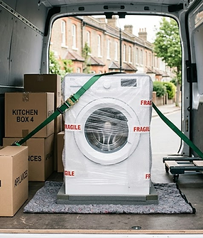 A secure washing machine wrapped in protective bubble wrap and fragile tape, anchored with a heavy-duty green tie-down strap inside the back of a white delivery van alongside organized kitchen appliance cardboard boxes on a South East London street.
