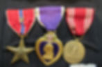 Three medals side by side pinned to pocket of military uniform. Left to right: Bronze Star Medal with "V" device, Purple Heart, Good Conduct Medal.
