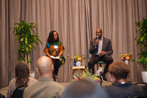 Two speakers sit on stage holding microphones and speak to an audience during a conversation.