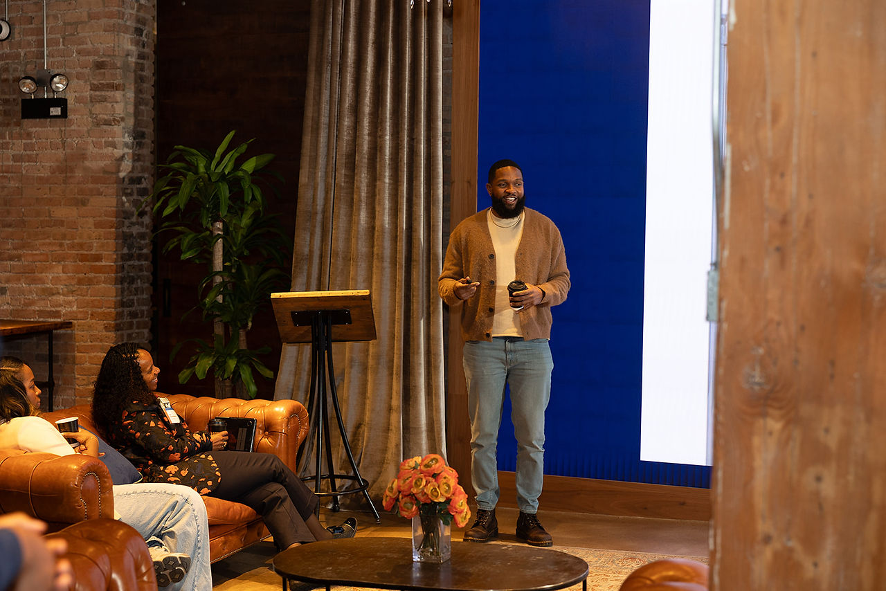 A speaker stands at the front of the room addressing seated attendees during a gathering.