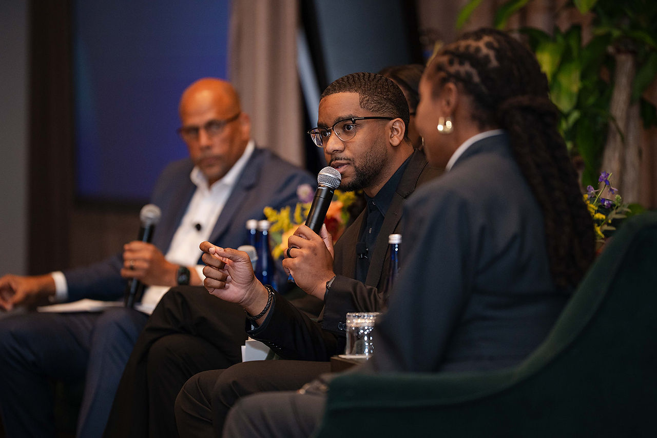 A panelist speaks into a microphone while seated on stage with other panelists during a discussion.