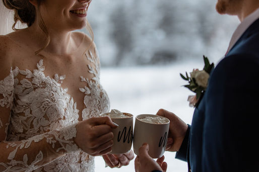 bride and groom having a hot drink in front of large glass window showcasing the snow covered scenery outside