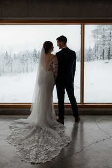 back view of bride and groom standing closely together in front of large window showing snow covered outside scenery