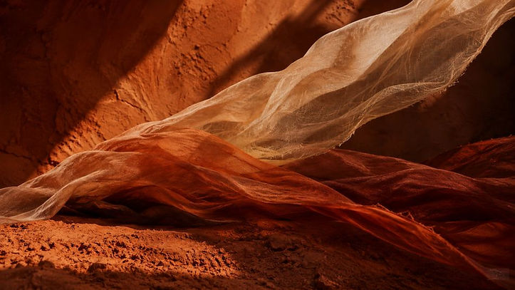Transparente Tücher in Terracotta- und Erdtönen lösen sich im Wind über einer rauen Landschaft.