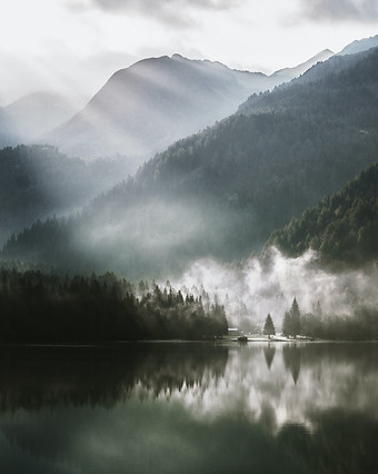 A golden arched window showing layers of forested mountains on a lake. The mountains have a light mist around them with pale sunlight streaming down through clouds. The mountains, mist, and light are reflected in a glassy body of water.