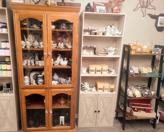 A display of vintage porcelain tea sets and decorative ceramics. The left side shows a wooden cabinet with glass doors containing various teacups, saucers, creamers, and swan figurines. The right side has open shelves with a large teapot, small tea cups, and more ceramic pieces. Some candle jars are visible on the far left.
