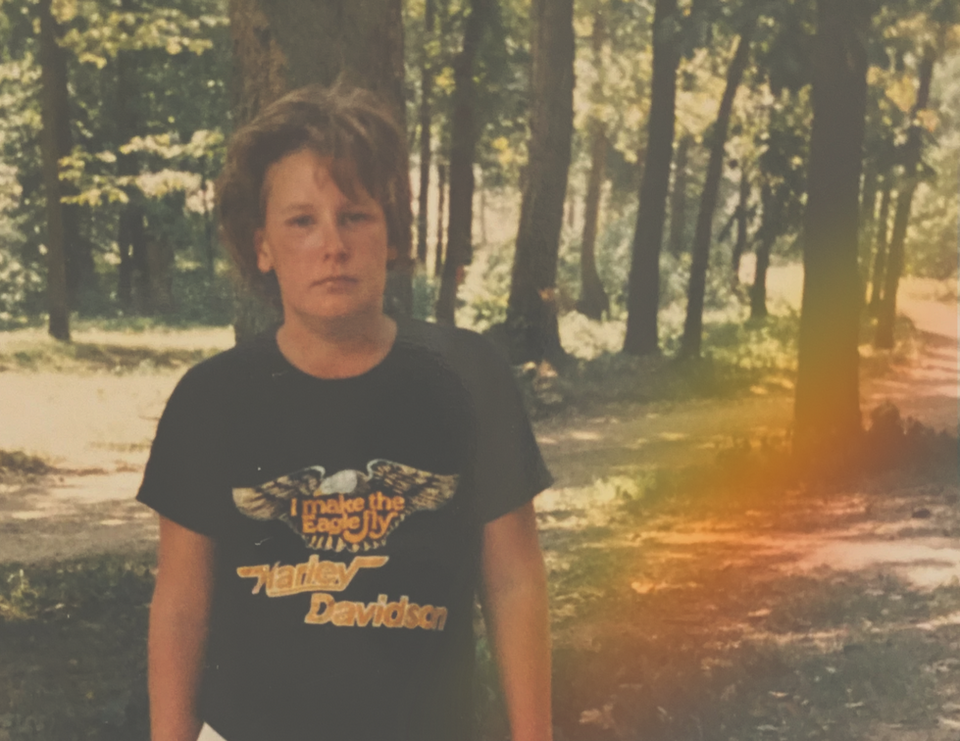 A dingy 80s teenager wearing a Harley Davidson t-shirt poses in a forest-filled campsite. He appears tired, as though he wasn't made for sleeping in a tent and that he's got much more important things to do. His t-shirt reads "I make the eagle fly" and a rainbow of light emerges to his left. 