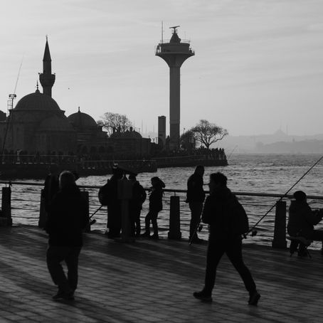Silhouettes of people fishing near a waterfront, with a mosque and tower in the hazy background. Monochrome image with a serene mood.