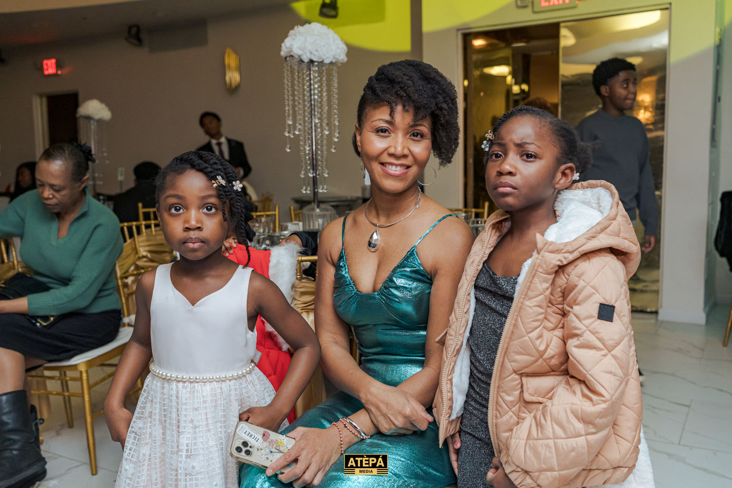 a woman in a green dress sits next to two little girls who are wearing white dresses