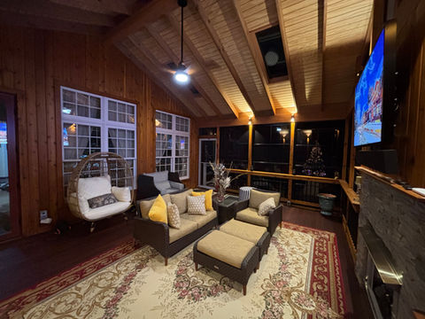 A cozy, wood-paneled screened porch at night featuring a vaulted wooden ceiling with exposed beams and a lit ceiling fan. The room is furnished with a wicker egg chair, a dark brown sofa with yellow accent pillows, and a large traditional floral rug over dark wood floors.