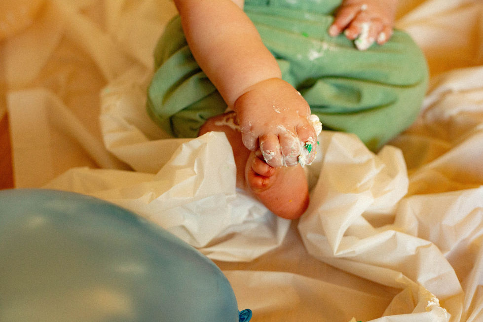 Young toddler wearing green overalls, holding his foot with icing covered fingers, edited in warm tones