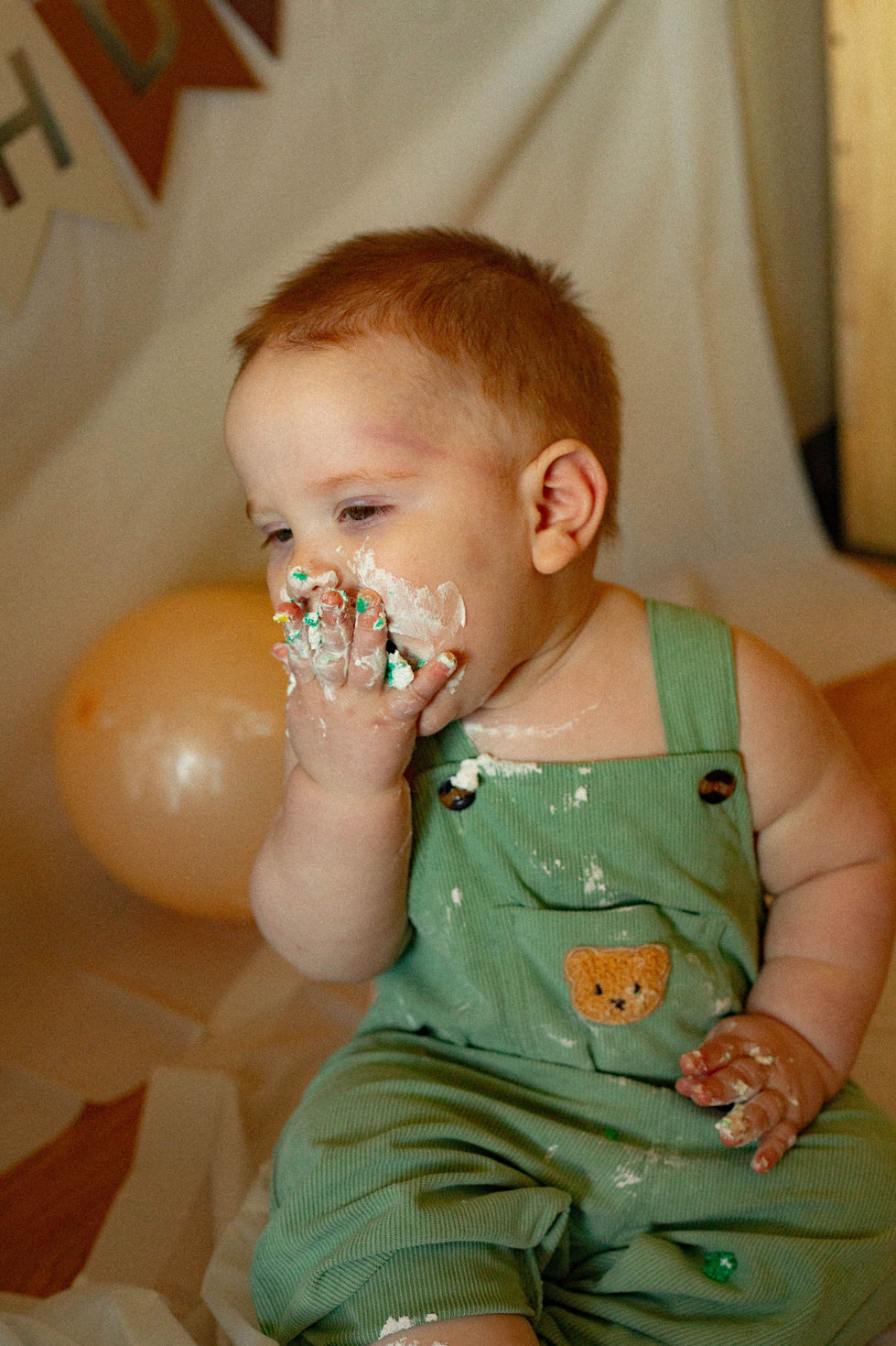 Young toddler wearing green overalls, eating icing off of his cake