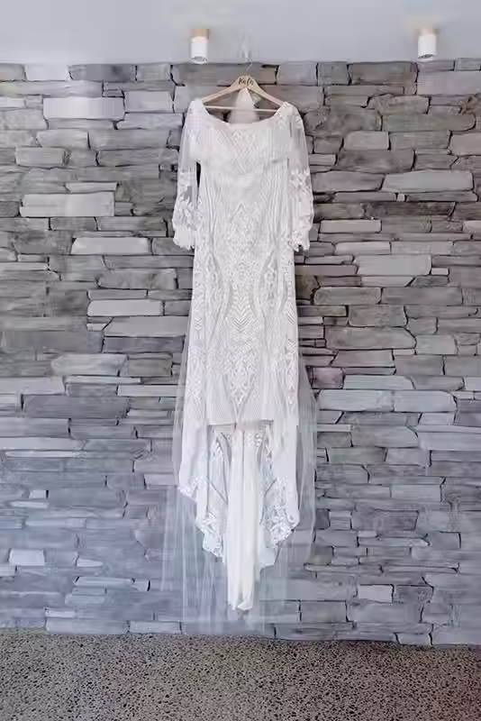 Bangor Farm wedding photography: white lace wedding dress with long sleeves and a flowing train hanging against a grey stone wall, Christchurch.