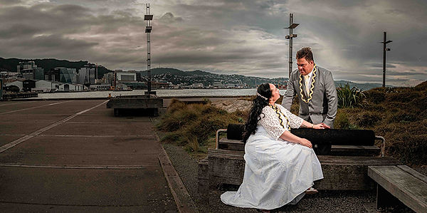 Auckland wedding photography and videography featuring a beautiful Māori couple sharing a heartfelt moment at the Wellington waterfront. Dressed in traditional wedding attire, the bride and groom are captured in a romantic pose against a dramatic cloudy sky and urban harbor backdrop. This stunning image blends cultural elegance with natural beauty, perfect for couples seeking meaningful, cinematic wedding portraits in New Zealand.