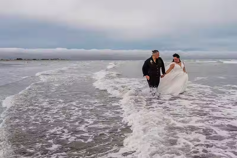 Christchurch Wedding Photography: Bride and groom walking through shallow waves on Sumner Beach. Captured by Christchurch Wedding Photographer Kiwi Moments Media.