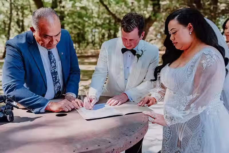 Bangor Farm wedding photography of the bride, groom, and an officiant signing marriage documents on a wooden table outdoors, a candid moment in Christchurch wedding.