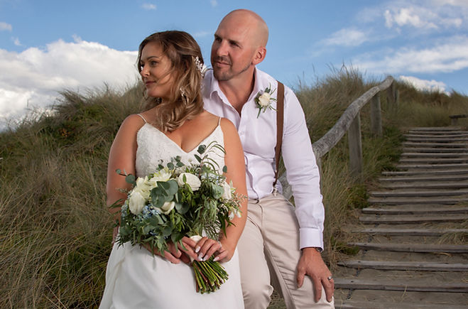 A wedding couple sits together overlooking the beautiful coastal nature landscape at New Brighton Beach, Christchurch, with the bride holding her bridal bouquet while they look out into the distance
