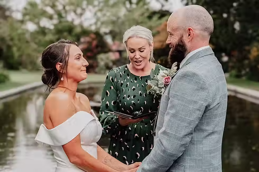 Bride and groom holding hands, looking at each other while an officiant conducts their outdoor wedding ceremony near a pond in Mona Vale, Christchurch.