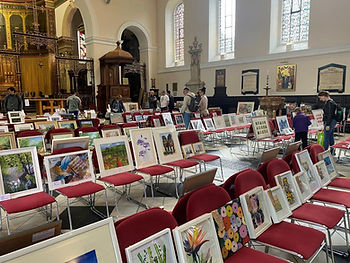 Art exhibition in a church hall, with framed paintings on red chairs as people browse beneath stained-glass windows.